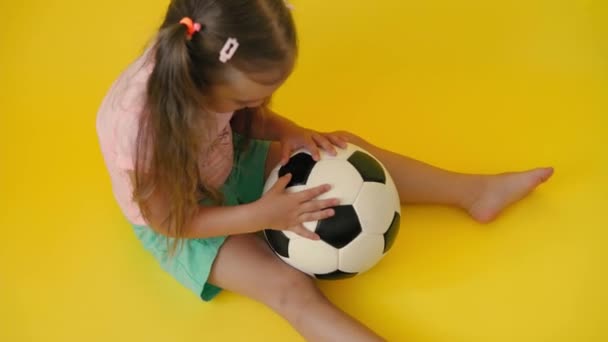 Authentique mignon souriant préscolaire petite fille avec ballon de football classique noir et blanc regarder la caméra sur fond jaune. enfant jouer au football en t-shirt et short. Sport, championnat, concept d'équipe — Video