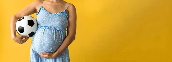 Motherhood, femininity, football, sport, dairy, hot summer. banner pregnant young pretty woman in floral blue dress holds soccer white and black leather classic ball rubs tummy on yellow background — Stock Photo, Image