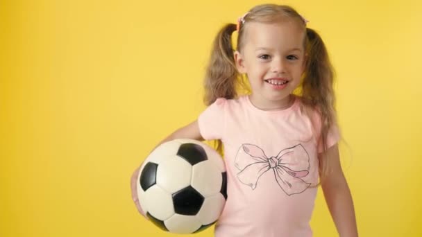 Αυθεντικό χαριτωμένο χαμογελαστό κοριτσάκι προσχολικής ηλικίας με κλασική ασπρόμαυρη μπάλα ποδοσφαίρου ματιά στην κάμερα σε κίτρινο φόντο. παιδί παίζει ποδόσφαιρο με t-shirt και σορτς. Αθλητισμός, πρωτάθλημα, ομαδική ιδέα — Αρχείο Βίντεο
