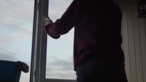 Lavando cristales de ventana en casa. Un joven de pelo largo con barba está limpiando el apartamento. El trabajador sanitario está desinfectando ventanas panorámicas en una habitación vacía. pureza, limpieza, cambio de concepto — Vídeos de Stock