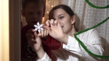 Otantik sevimli bir anne ve iki küçük anaokulu öğrencisi kız ve erkek kardeş 2-4 yaşlarında pencereye Noel ağacı çiziyor. Çocuklu genç bir kadın kar taneleri yapıştırıyor. Xmas, yeni yıl, kış konsepti.