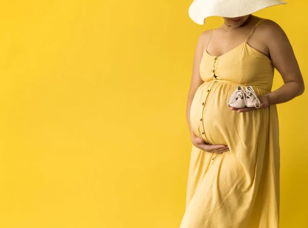Maternidade, feminilidade, moda, verão quente - banner Close-up mulher grávida irreconhecível em belo vestido vintage Provence chapéu de palha com pequenos sapatos de bebê no fundo amarelo, espaço de cópia — Fotografia de Stock