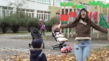 Çocukluk, aile, bebek, annelik, sonbahar konsepti - yavaş çekim 2 mutlu küçük anaokulu çocukları anneleriyle birlikte soğuk mevsimde oyun parkının yakınına bir sürü sarı yaprakları toplayıp atıyorlar.