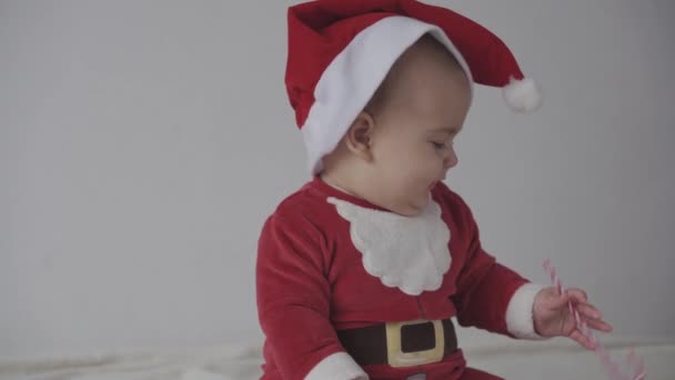 Feliz Navidad, feliz año nuevo, bebés, infancia, concepto de vacaciones - primer plano sonriendo 7 meses divertido bebé recién nacido en sombrero de Santa Claus, juego de body rojo sentado en la cama suave caliente lamer dulces dulces de Navidad — Vídeos de Stock