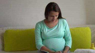 Not Defteri Hazırlama çantalarıyla doğum hastanesi için paketleme listesi hazırlayan sevimli hamile kadın. Hamilelikteki genç Ledy 'nin evde koltukta keyfi yerinde. Annelik, Sağlık ve Bakım Konsepti