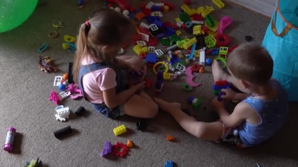 Kleine kleuters en broertjes en zusjes zitten op de vloer met speelgoed in de kinderkamer. De jongens spelen met de bouwvakker in de speelkamer. Kinderen hebben veel plezier thuis gehad. Jeugd, vriendschap, ontwikkelingsconcept — Stockvideo