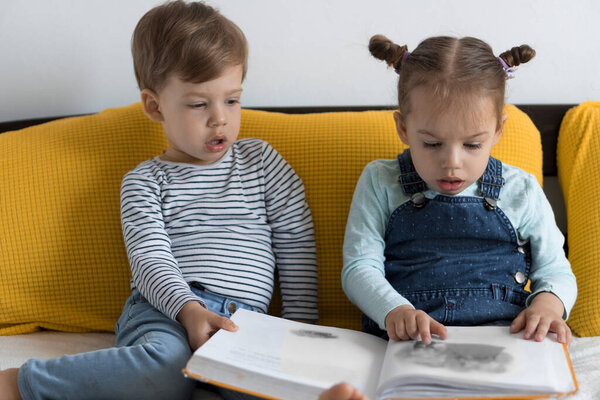 Two preshool, toddler children read large interesting book of fairy tales on yellow bed. Siblings little twins reader have fun, happy kid on quarantine at home. Friendship, family, education concept