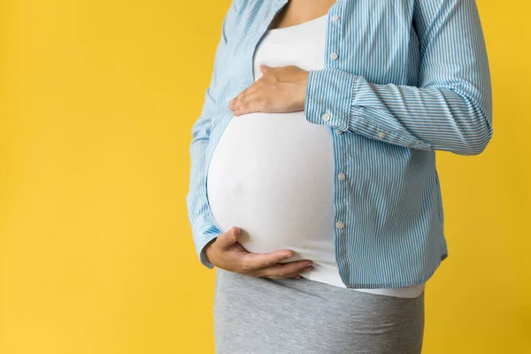 Maternidade, feminilidade, amor, cuidado, espera, verão quente - brilhante cortado Close-up mulher grávida irreconhecível na camisa com sapatos pequenos bebê mão sobre barriga esfregar barriga no fundo amarelo, espaço cópia — Fotografia de Stock