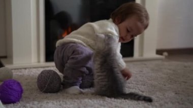 Bebek kediyle oynuyor. Erkek kedi halının üstünde, şöminenin yanında, konforunda. Çizgili kedi yavrusu iplik yumağıyla oynuyor. Kitty kameraya bakarak koş. Mutlu sevimli hayvan, çocukluk, vahşi doğa konsepti