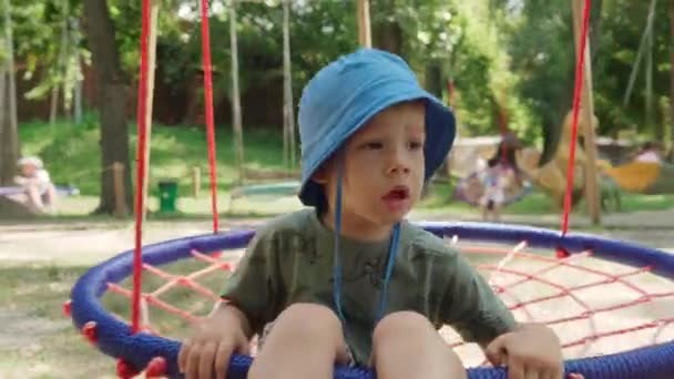 Little Cute Prechool Baby Boy in Panama Hat Ride on Hanging Hammock Swing on Playground in Public Park. Дети весело проводят время в летний солнечный полдень на открытом воздухе. Детство, концепция развития — стоковое видео