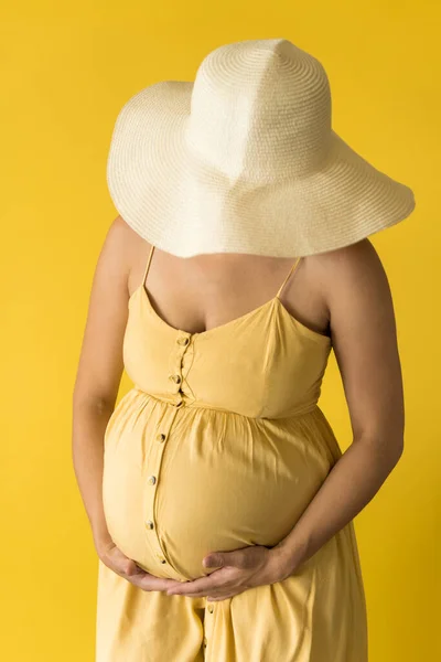 Maternidade, feminilidade, moda, verão quente - cortada Close-up mulher grávida irreconhecível em belo vestido vintage Provence chapéu de palha com as mãos sobre a barriga esfrega barriga no fundo amarelo — Fotografia de Stock