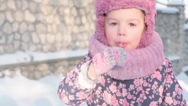 Zima, wakacje, gry, koncepcje rodzinne - slo-mo zbliżenie autentyczne małe przedszkole moll 3-4 lat dziewczyna w fioletowym patrząc na aparat liże sopl z języka na śniegu. dziecko biegnie po śnieżnej okolicy — Wideo stockowe