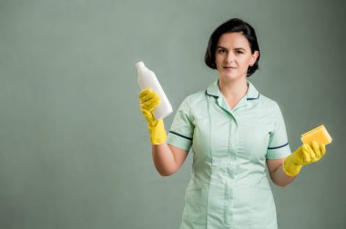 Yeşil gömlekli ve sarı eldivenli genç temizlikçi kadın, elinde bulaşık çözeltisi ve yeşil arka planda izole edilmiş sünger tutuyordu.