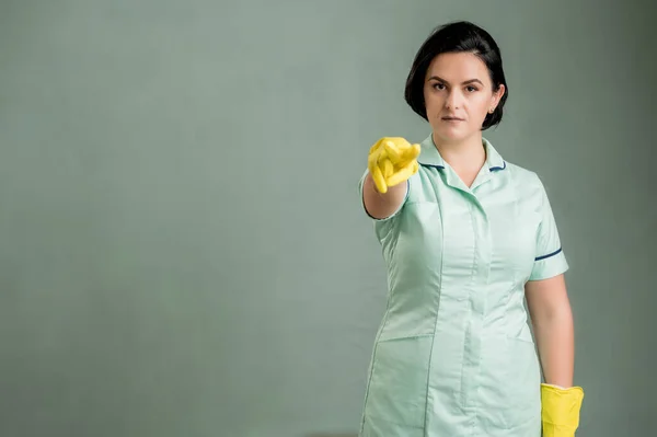 Young cleaning woman wearing a green shirt and yellow gloves, doing I am watching you gesture isolated on green background