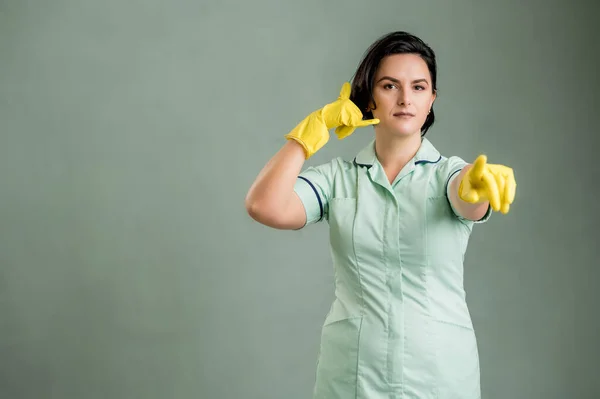 Young cleaning woman wearing a green shirt and yellow gloves, showing call me gesture, pointing her finger isolated on green background