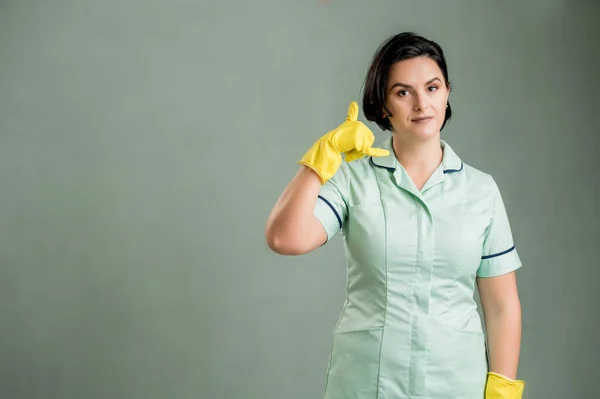 Young cleaning woman wearing a green shirt and yellow gloves, showing call me gesture isolated on green background