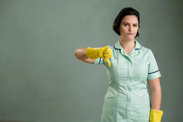 穿着绿色衬衫和黄色手套的年轻清洁女工 在绿色背景下显得很不喜欢 图库图片
