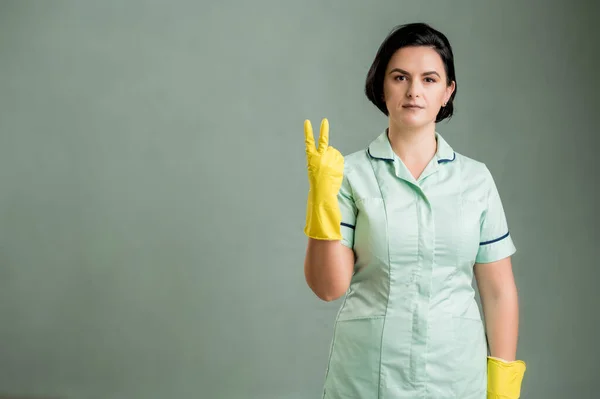 穿着绿色衬衫和黄色手套的年轻清洁女工 手指被绿色背景隔开 数着两个 图库照片