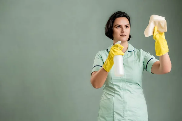 穿着绿色衬衫和黄色手套的年轻清洁妇女 手握窗户清洁液 与绿色背景隔离 图库图片