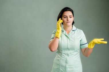 Yeşil gömlekli ve sarı eldivenli genç temizlikçi kadın, hoşnutsuz yüz ve jestler ve yeşil arka planda izole edilmiş cep telefonuyla konuşan.