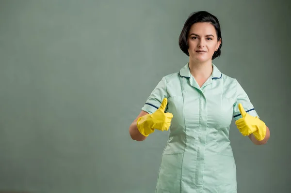 穿着绿色衬衫和黄色手套的年轻清洁女工 双手被绿色背景隔离 竖起大拇指 免版税图库图片