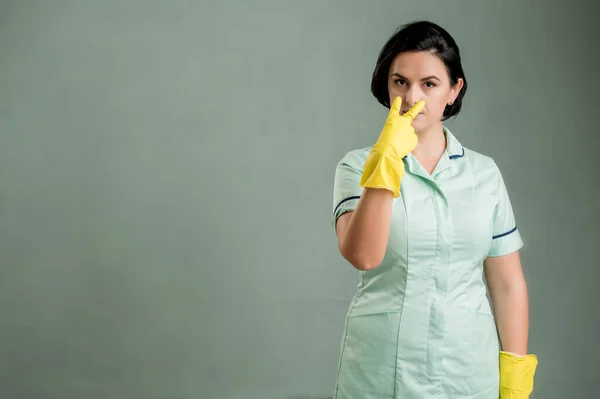 Young cleaning woman wearing a green shirt and yellow gloves, making see me gesture isolated on green background