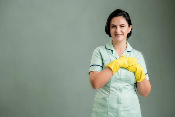 Young cleaning woman wearing a green shirt and yellow gloves, holding hands near chest isolated on green background