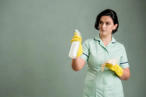 Young cleaning woman wearing a green shirt and yellow gloves, holding window cleaning solution in hands isolated on green background