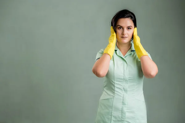 穿着绿色衬衫和黄色手套的年轻清洁女工 耳朵像聋子一样被隔离在绿色背景中 免版税图库照片