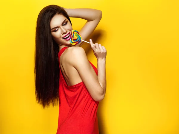 Bella ragazza con caramelle nelle mani sorridente e in posa, T-shirt rossa, capelli belli e sani, sfondo giallo — Foto Stock