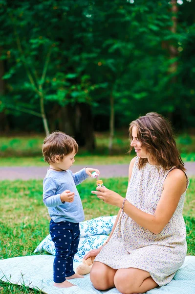妊娠中、幸せな母親と一緒に芝生の夏に公園でシャボン玉を吹く少年 — ストック写真