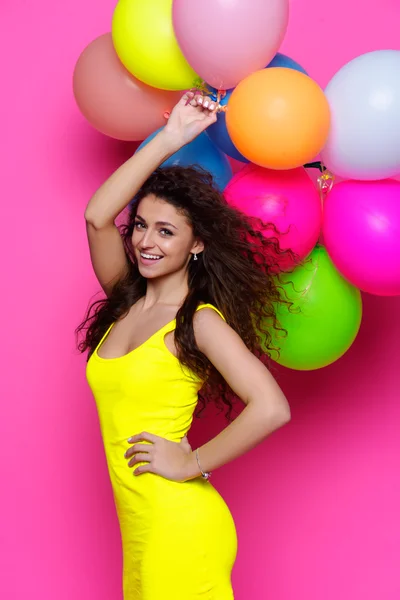 Unga och vackra lockiga flicka i en gul klänning på rosa bakgrund håller färgglada ballonger — Stockfoto