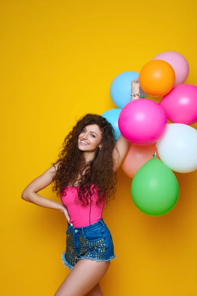 Jong en mooi krullend meisje in een roze shirt en blauwe korte broek op een gele achtergrond houdt van kleurrijke ballonnen en lachen — Stockfoto