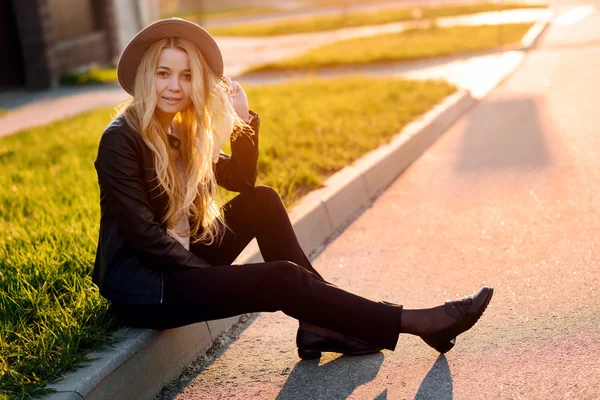Ung och vacker flicka i en hatt som sitter på trottoarkanten sida av vägen vid solnedgången — Stockfoto