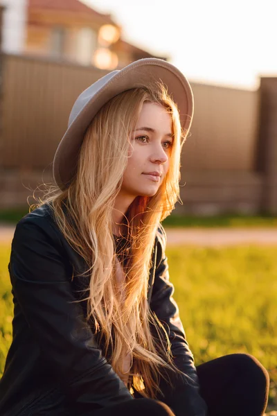 Młoda i piękna dziewczyna w kapeluszu siedzi na stronie krawężnik drogi na zachodzie słońca — Zdjęcie stockowe