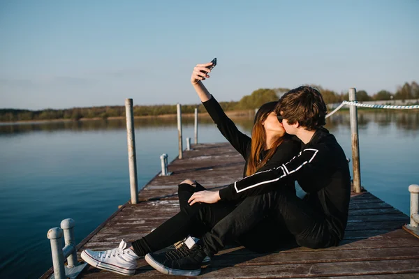 Joven pareja feliz en gafas de sol abrazándose y besándose en un muelle fotografiado por teléfono en un día soleado de verano. Concepto de amor —  Fotos de Stock