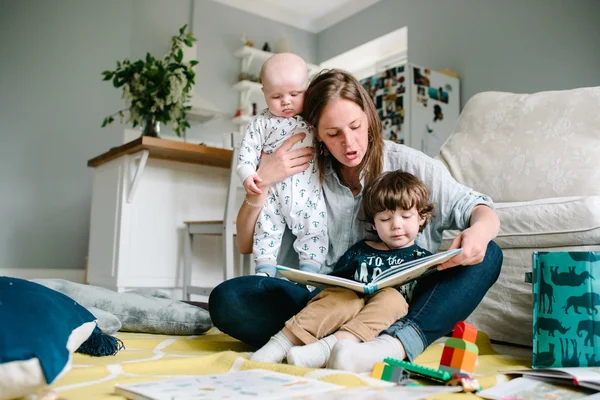 Lycklig ung mamma läser en bok till sina barn sitter i golvet i huset. Begreppet familje — Stockfoto