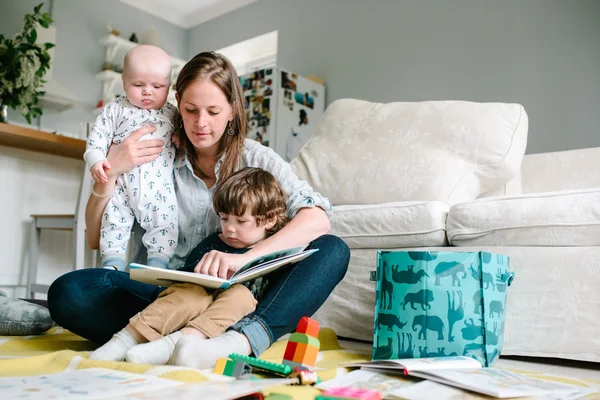 Lycklig ung mamma läser en bok till sina barn sitter i golvet i huset. Begreppet familje — Stockfoto