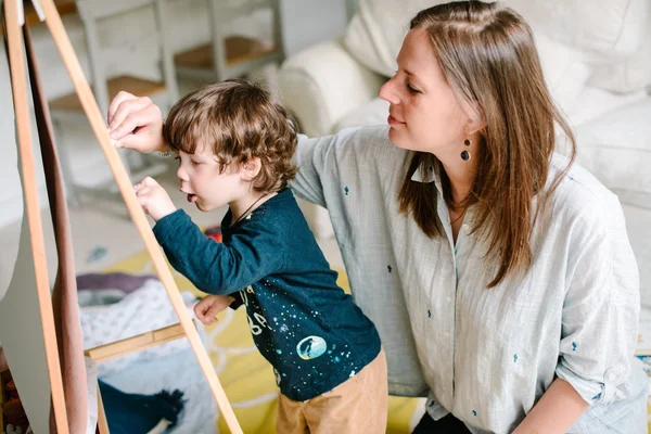 La jeune mère dessine une craie sur un tableau noir avec son jeune fils. Le concept de famille — Photo