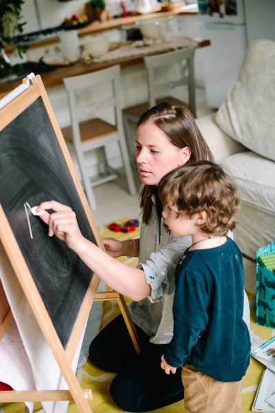 La joven madre dibuja una tiza en una pizarra con su hijo pequeño. El concepto de familia — Foto de Stock