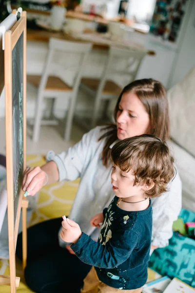 La jeune mère dessine une craie sur un tableau noir avec son jeune fils. Le concept de famille — Photo