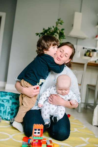 Mère jeune et heureuse embrassant leurs jeunes enfants sur le sol à la maison. Les enfants rient. Concept de famille. Fils embrasse mère — Photo