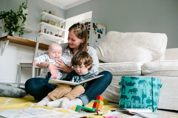 Feliz joven madre leyendo un libro a sus hijos sentados en el suelo de la casa. El concepto de familia — Foto de Stock