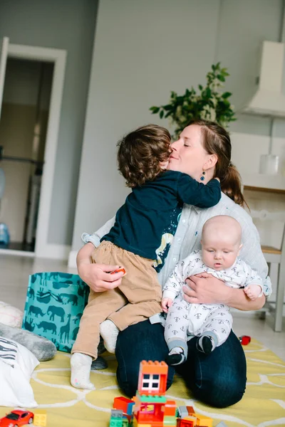 Mladá a šťastná matka objímající své malé děti doma na podlaze. Děti se smějí. Pojem rodina. Syn líbá matku — Stock fotografie