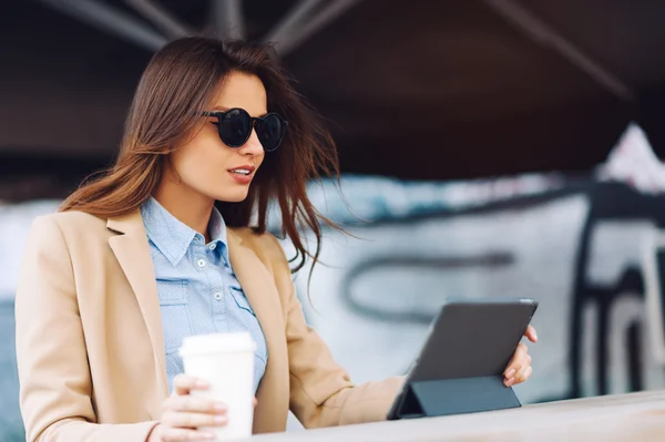 Güzel ve genç kız sokakta kahve içme ve bir Tablet haber okuma güneş gözlüğü — Stok fotoğraf