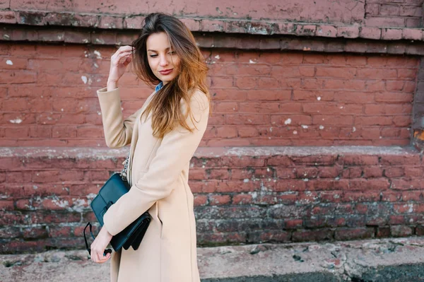 Hermosa y joven chica en abrigo de moda y hermoso pelo largo caminando en la ciudad de verano. Sobre el fondo de la pared de ladrillo rojo . — Foto de Stock