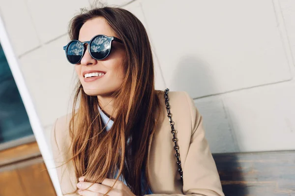 Hermosa y joven chica en un abrigo y bufanda y gafas de sol sentado en el banco y se ríe. Verano. El sol — Foto de Stock