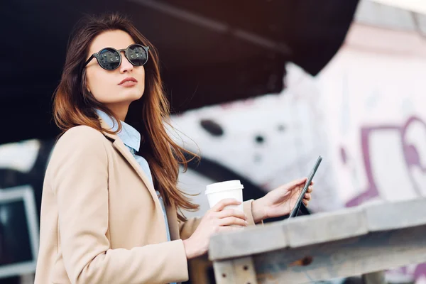 Hermosa y joven chica en gafas de sol tomando café en la calle y leyendo las noticias en una tableta — Foto de Stock