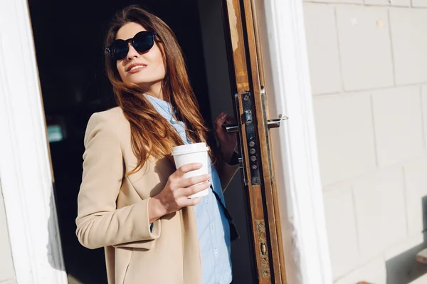 Hermosa y joven chica saliendo de un café y sosteniendo una taza de café en gafas de sol . —  Fotos de Stock