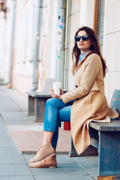 Hermosa y joven chica en un abrigo y bufanda y gafas de sol sentado en el banco. Mujer bebiendo café. Verano . —  Fotos de Stock